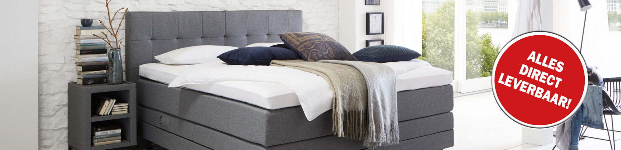 Grijze boxspring met witte beddengoedlagen en decoratieve kussens, voorzien van een rode sticker 'alles direct leverbaar!'