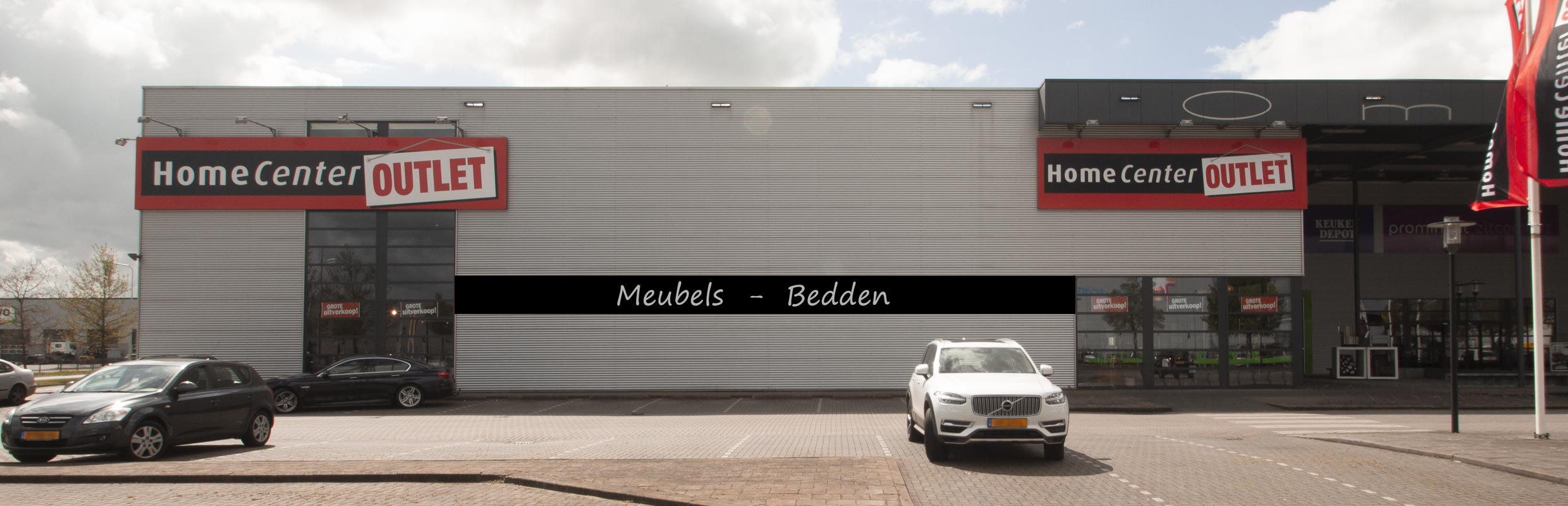Vooraanzicht van Home Center Outlet, met een groot rood-wit bord en de tekst 'Meubels - Bedden ' op het gebouw.